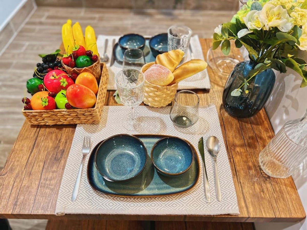 A wooden table set with blue plates, a basket of fruits, bread, and a vase with flowers.