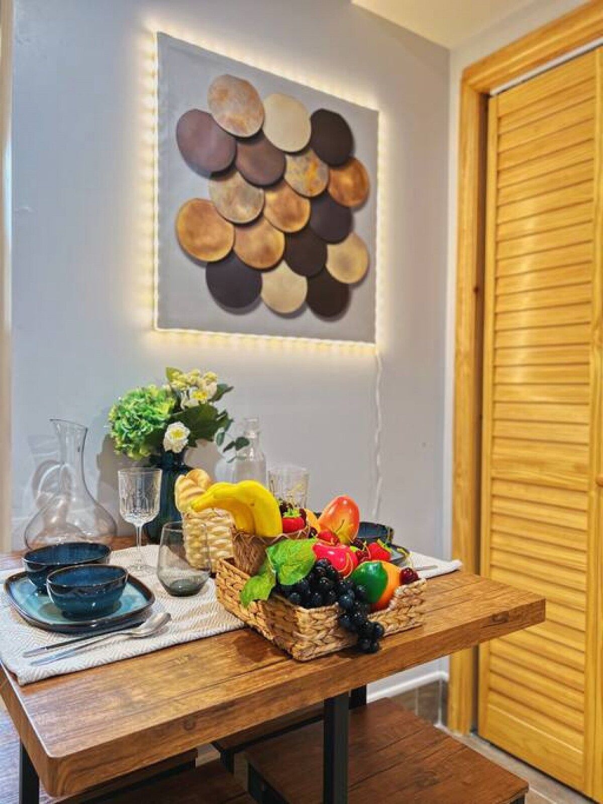 A wooden table with a fruit basket, a vase with flowers, and a wall-mounted decorative piece.