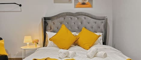 A bed with a tufted headboard, two pillows, a rolled white towel, and a yellow blanket.