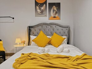 Un lit avec une tête de lit capitonnée, deux oreillers, une serviette blanche roulée et une couverture jaune.