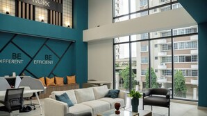 A modern living room with a sofa, armchairs, and a coffee table. There is a desk with a chair and a computer. The room has large windows and a view of buildings outside.