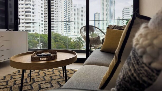 A modern living room with a sofa, a round wooden coffee table, and a chair. A television is mounted on the wall. The room has large windows offering a view of high-rise buildings and greenery outside.
