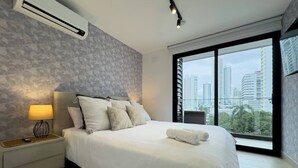A modern hotel room with a large bed, a nightstand, a lamp, and a city view through a large window.