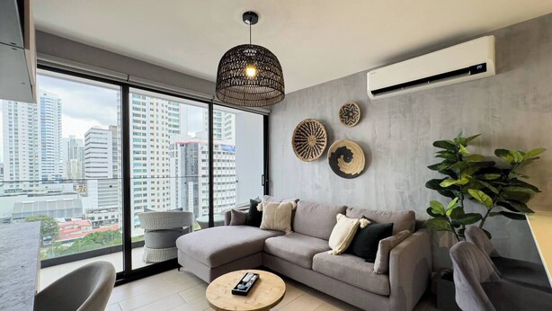A modern living room with a large window, a grey sectional sofa, a round wooden coffee table, and a hanging woven light fixture. - Modern and cozy apartment in the heart of Panama City, ideal for business travelers and digital nomads. Pool, gym, and coworking space available, with valet parking offered as an optional service. (Panamá)