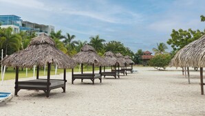 Una playa con sombrillas de paja, bancos de madera, palmeras y un edificio moderno al fondo.