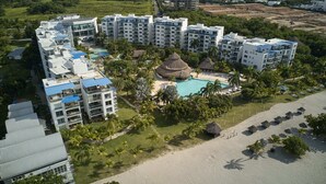 Un resort con varios edificios altos, un área de piscina central con una estructura de techo de paja y una playa con tumbonas.