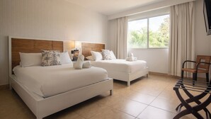 Habitación de hotel con dos camas, cabecera de madera, mesita de noche con lámpara y vista a zonas verdes por la ventana.