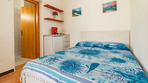 A bedroom with a bed, a wooden door, a small desk with a red object, and two wall-mounted shelves with items. - Villa Bouganville   Il Gelsomino (CAPOLIVERI)