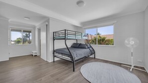 Een kamer met een stapelbed, een ventilator, een klein wit tafeltje en een groot raam met uitzicht op buiten.