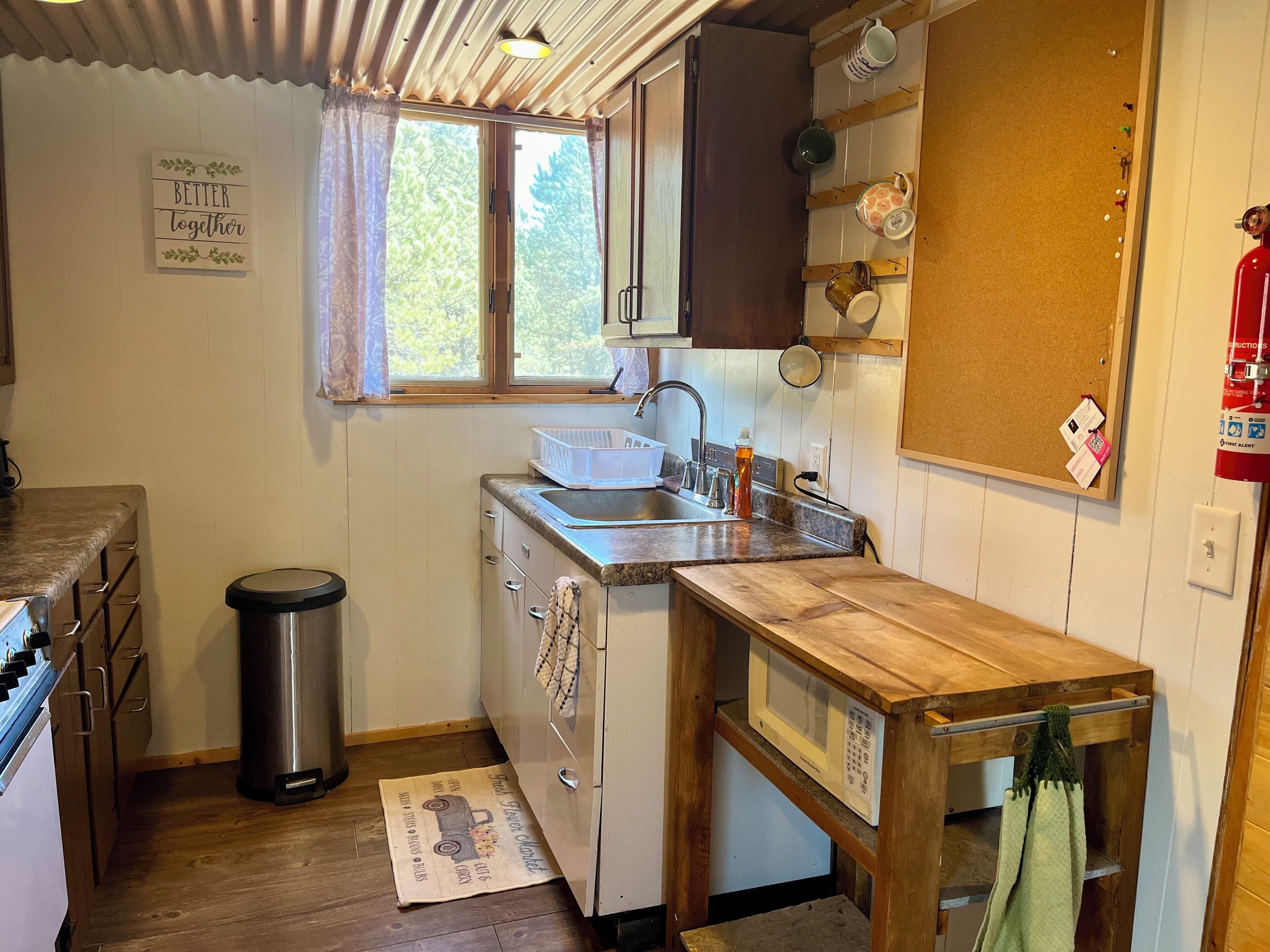 Une cuisine compacte équipée d’un évier, d’une cuisinière et d’un four à micro-ondes. On y trouve une fenêtre avec des rideaux, un tableau d’affichage en liège et une petite poubelle.