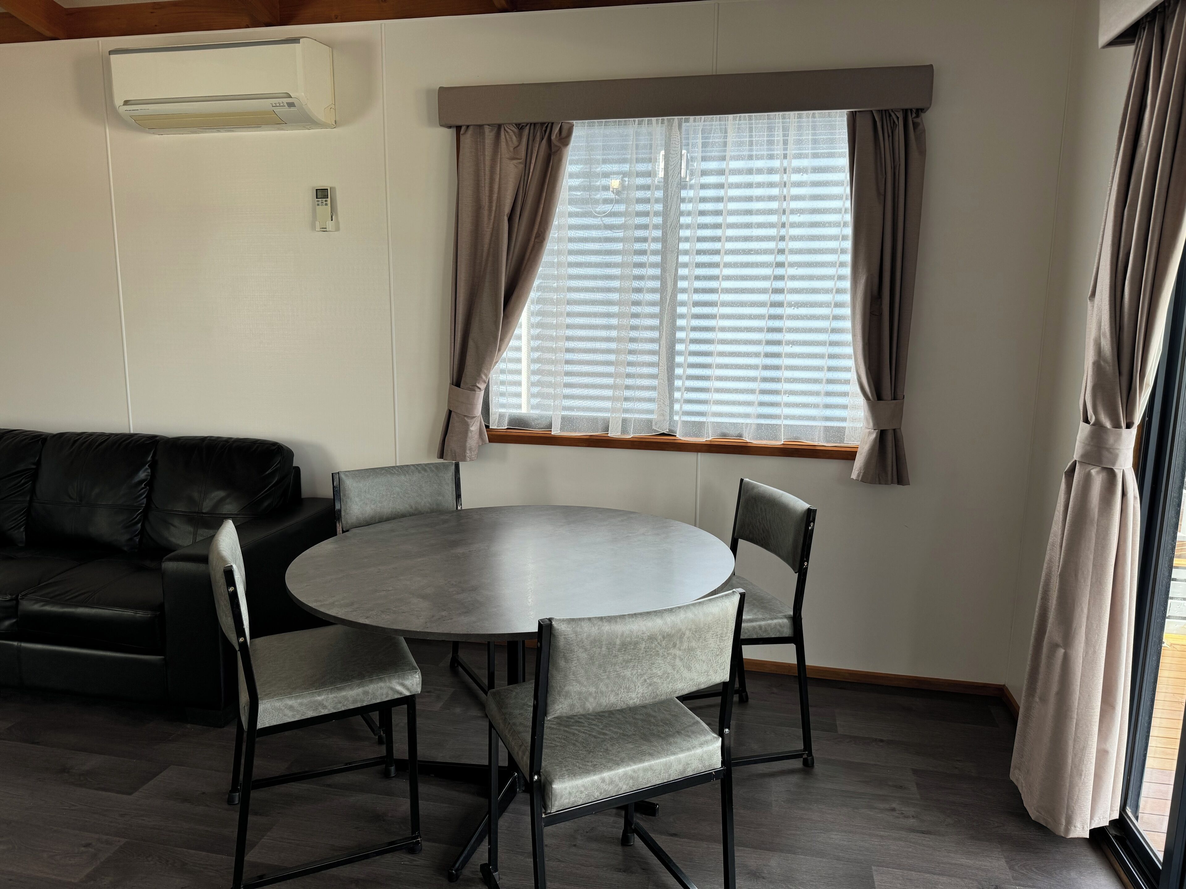 Een kamer met een ronde tafel, vier stoelen, een zwarte leren bank en een raam met gordijnen.