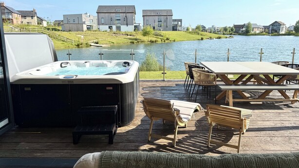 Una vasca idromassaggio su un pontile di legno con vista su un lago e case sullo sfondo.