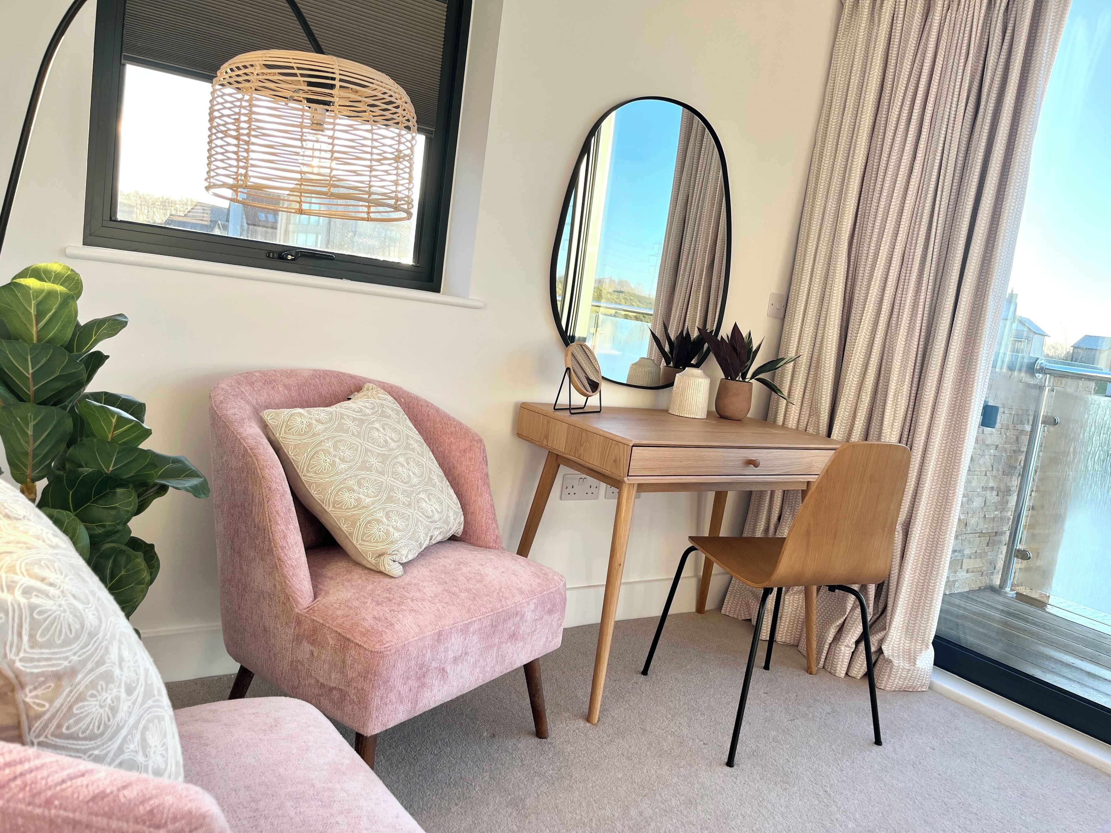 Une chambre moderne avec un fauteuil rose, un bureau en bois, un miroir et une grande fenêtre avec des rideaux.
