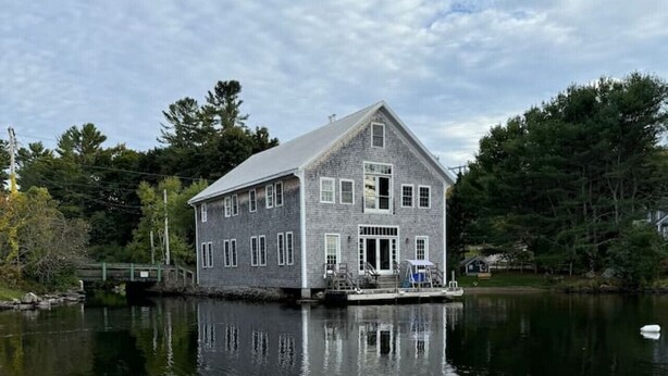 A large, single-story house with a prominent front door and windows, situated by a calm lake with a bridge in the background. - More Boathouse than Lakehouse (Newcastle)