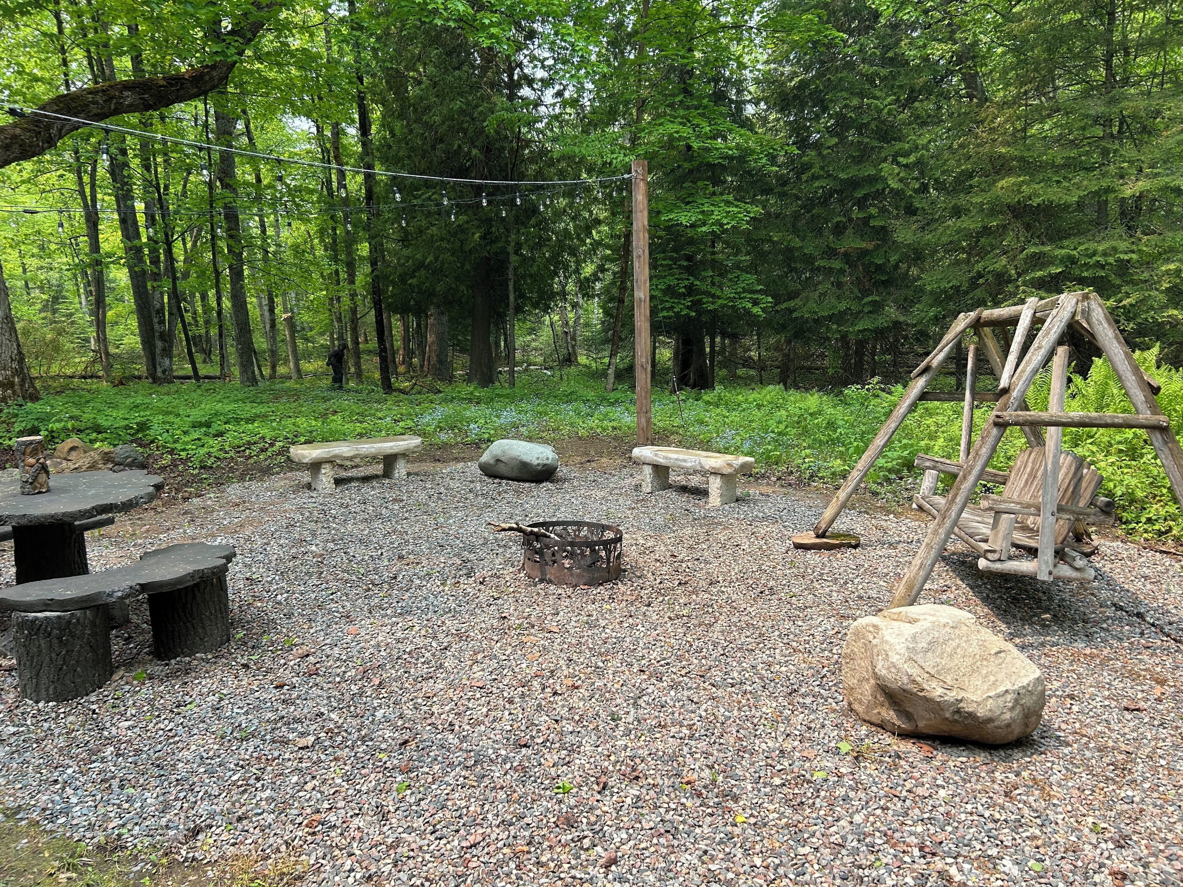 A forested area with a wooden swing set, stone benches, and a fire pit.