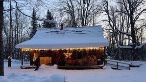 Une cabane dont le toit est recouvert de neige, une véranda décorée d’une étoile et une pile de bois de chauffage à l’extérieur.