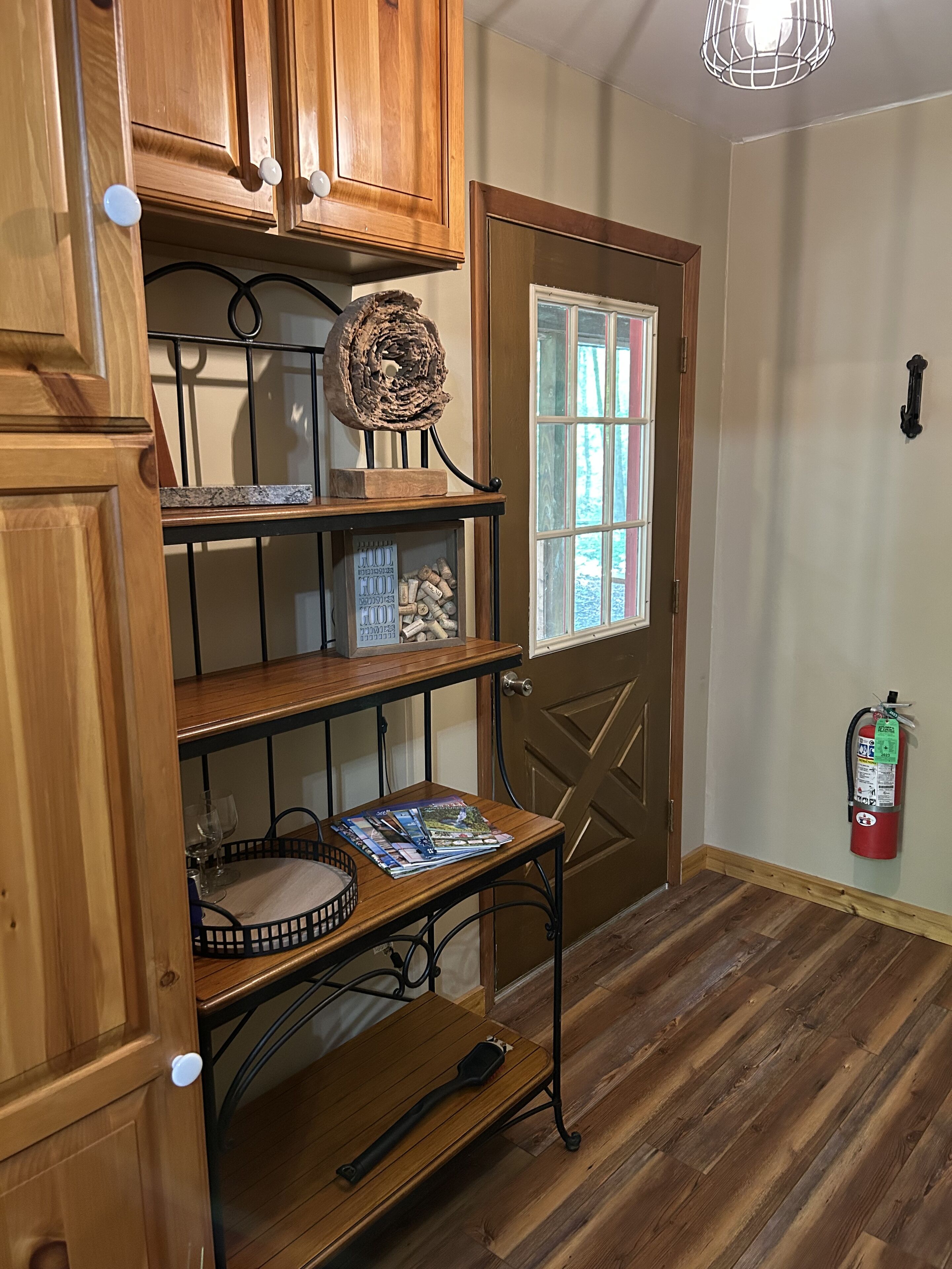 Interior de madera con una puerta, una estantería con copas de vino, un extintor y una ventana con persianas.