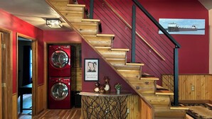 Un escalier en bois, un mur rouge et un tableau représentant un navire. On y trouve également deux machines à laver rouges et une petite table ornée d’objets décoratifs.