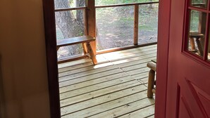 Une terrasse en bois avec un banc, une porte rouge et une vue sur une forêt, visible à travers de grandes fenêtres.