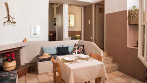 Un comedor con una mesa para dos, un banco con cojines, una chimenea con un cesto de flores y una escalera que lleva a otra habitación.