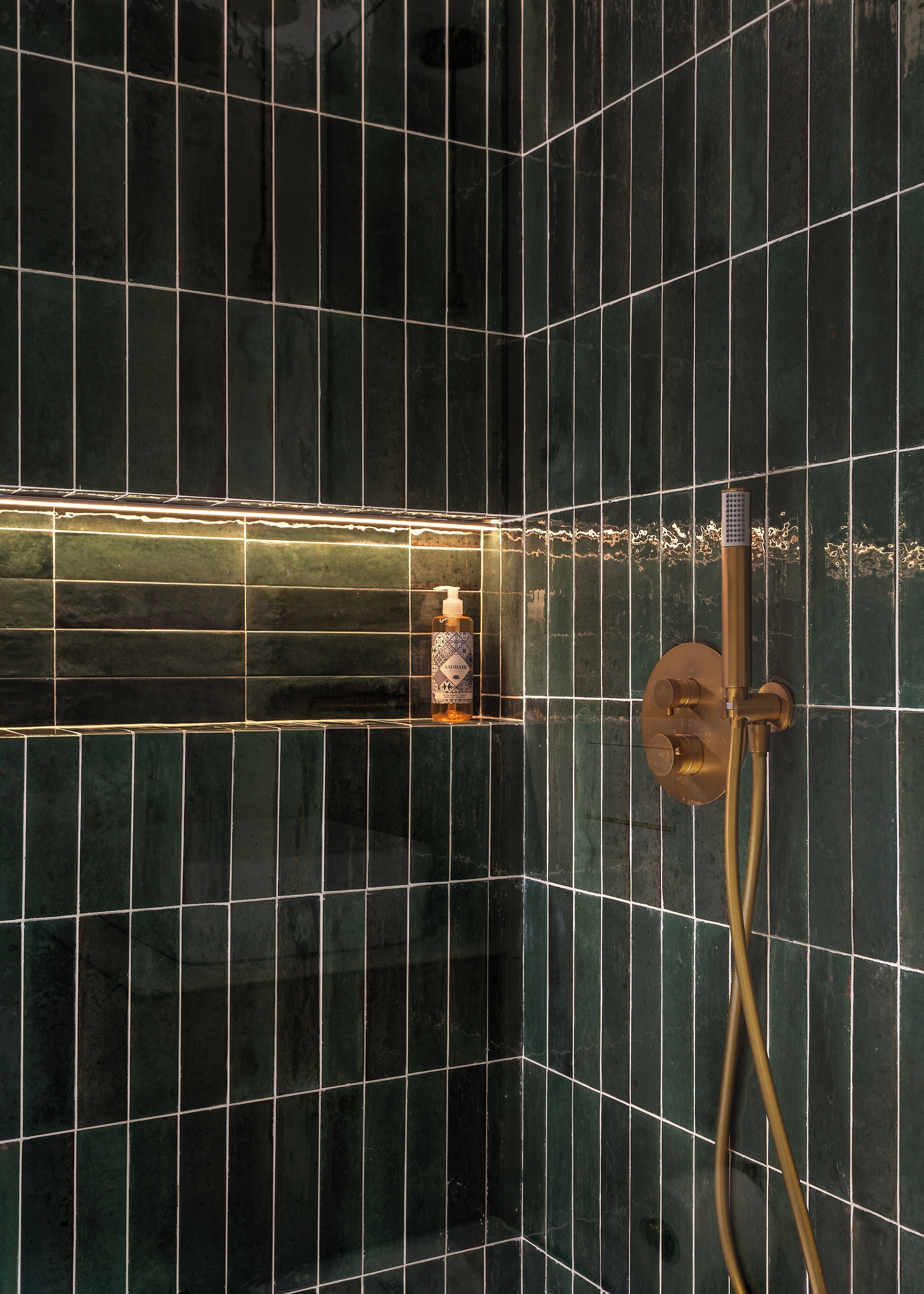 A bathroom with dark green tiles, a handheld showerhead, and a bottle of hand soap.