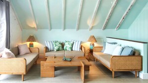 A cozy room with a high ceiling, wooden beams, and a light blue wall. There are two wicker sofas with cushions, a wooden coffee table, and two lamps.