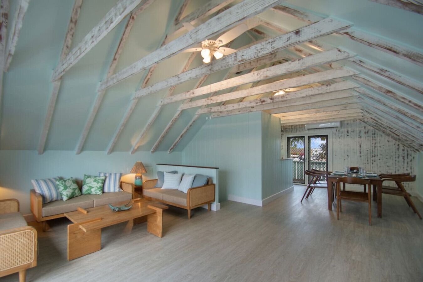 A spacious attic room with exposed wooden beams, a sofa set, a dining table, and a view through a glass door.