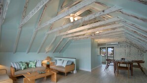 A spacious attic room with exposed wooden beams, a sofa set, a dining table, and a view through a glass door.