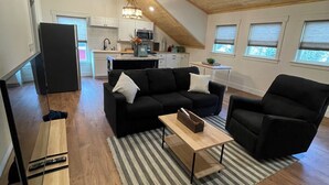 A spacious living area with a black sofa, a coffee table, and a TV stand. A kitchen is visible in the background with a ceiling fan and windows.