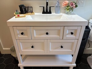 Un tocador de baño blanco con lavamanos, varios cajones y una encimera con artículos de aseo.