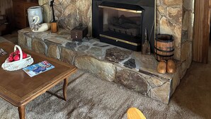 A stone fireplace with a black surround, a wooden coffee table with magazines, and a rustic wooden shelf.