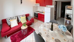 A compact living space with a kitchenette, a red sofa with patterned cushions, a dining table with a floral tablecloth, and a small side table with a plant. - Amazing apartment in Supetar with lake view (Supetar)