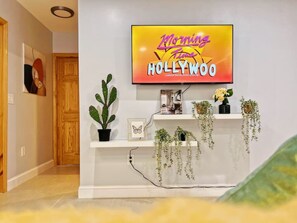 A living room with a flat-screen TV displaying 'Morning Time Hollywood,' a cactus, and shelves with plants and books. - Ramblei near Chinatown & CenterCity | 1Bedroom-B (Philadelphia)