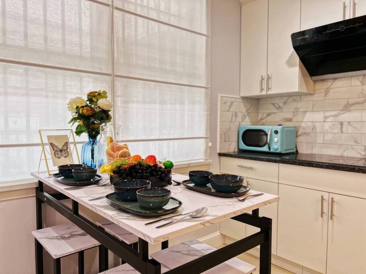 Een keuken met een marmeren aanrecht, een blauwgroene magnetron en een eettafel gedekt met kommen en borden.