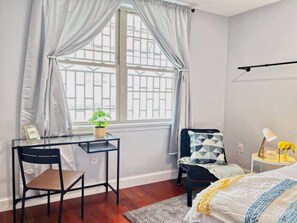 A bedroom with a bed, a desk with a chair, a window with curtains, a bedside table with a lamp, and a small plant.
