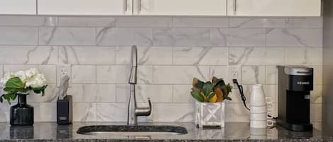 A kitchen with a marble backsplash, a stainless steel faucet, a coffee maker, and a vase with flowers.