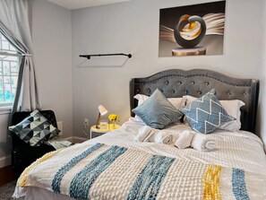 A neatly made bed with a striped blanket, a bedside table with a lamp and flowers, and a wall-mounted artwork. - Ramblei near Chinatown & CenterCity | 1Bedroom-B (Philadelphia)