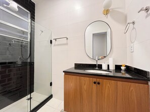 A modern bathroom with a glass shower enclosure, a wooden vanity with a black countertop, and a large mirror above the sink. - Toque a Campainha 207 Vendaval (Cabo Frio)