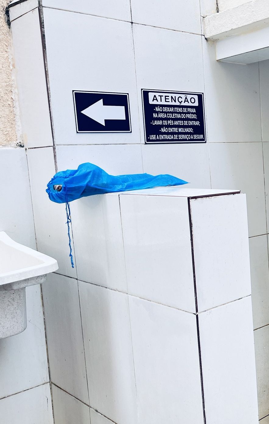 Ein Badezimmer mit weiß gefliesten Wänden, einer blauen Plastiktüte, die an einem Haken hängt, und einem Schild mit einem Pfeil und Text auf Portugiesisch.