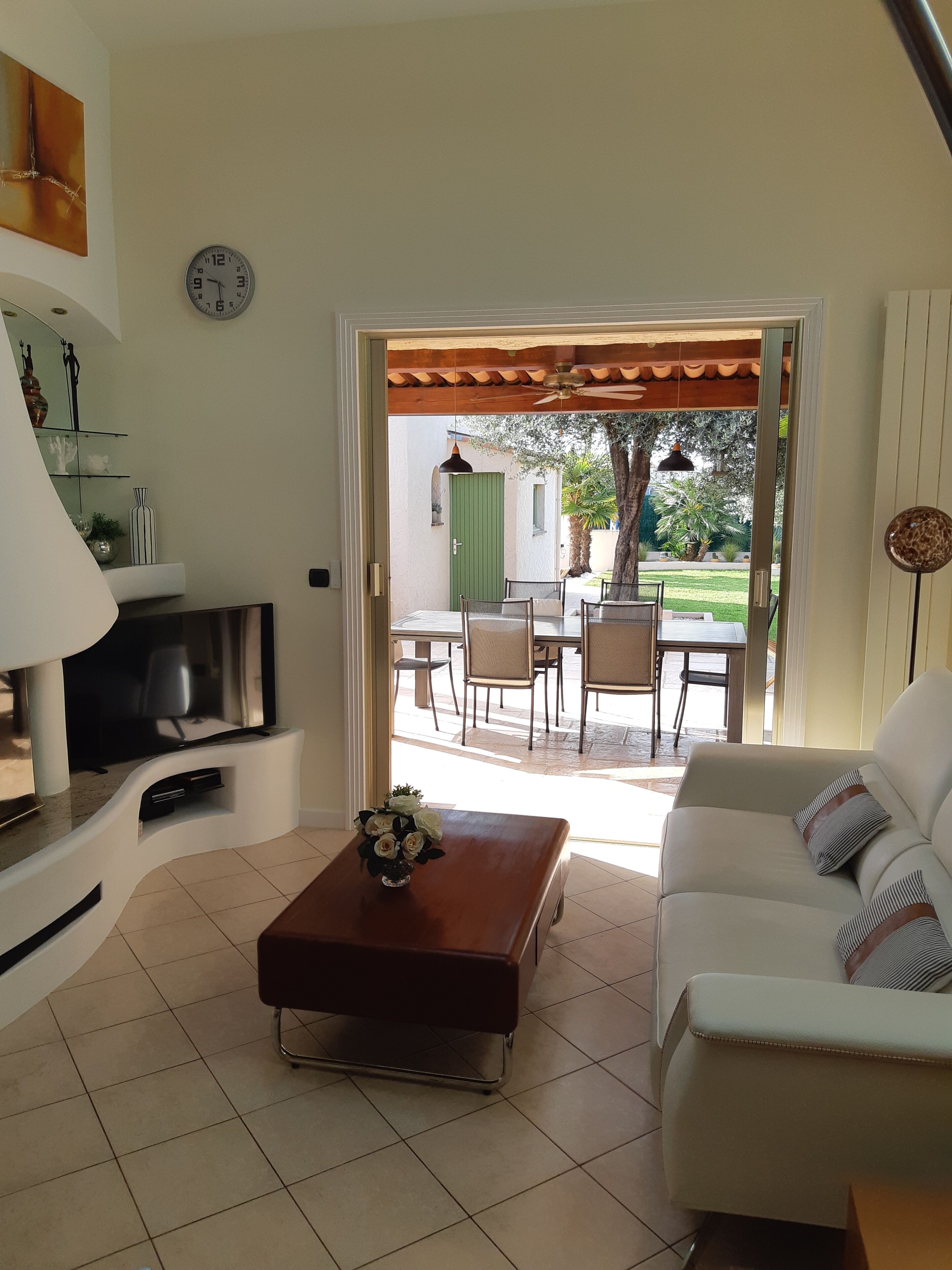 Un salon avec un téléviseur, une table basse ornée de fleurs, un canapé et une porte ouverte donnant sur une terrasse extérieure avec une table et des chaises.