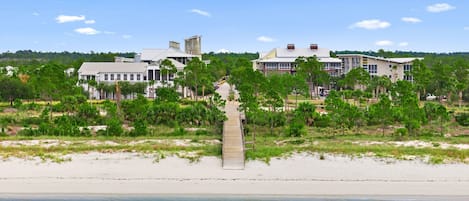 Une propriété située en bord de mer, avec un bâtiment blanc imposant, une promenade en bois menant à la plage et une végétation luxuriante.