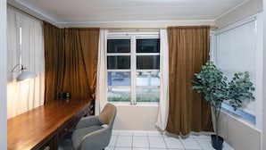 Et værelse med et træskrivebord, en stol, et vindue med gardiner, en potteplante og en lampe.
