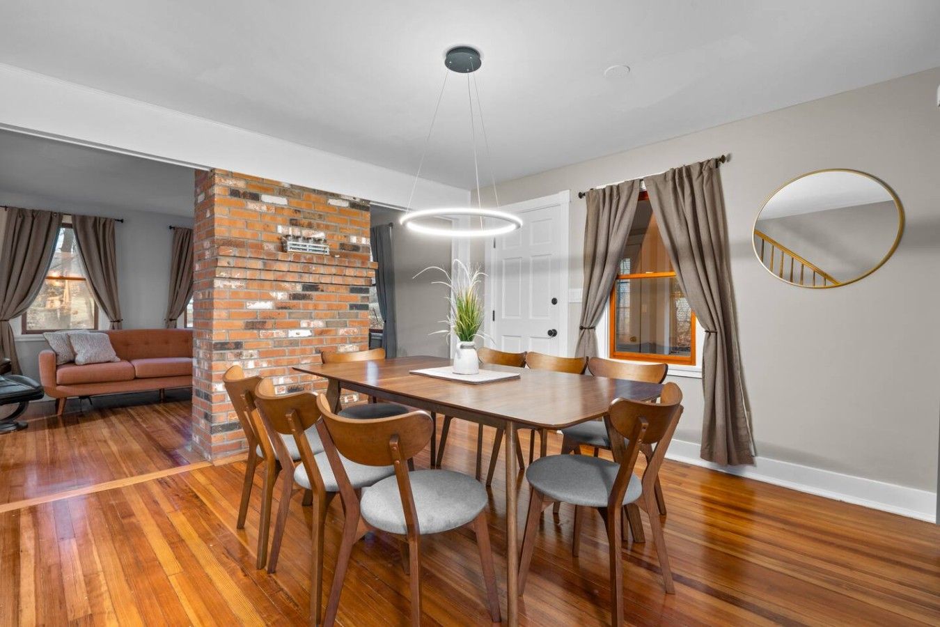 A modern dining area with a wooden table, six chairs, a hanging light fixture, and a brick accent wall.