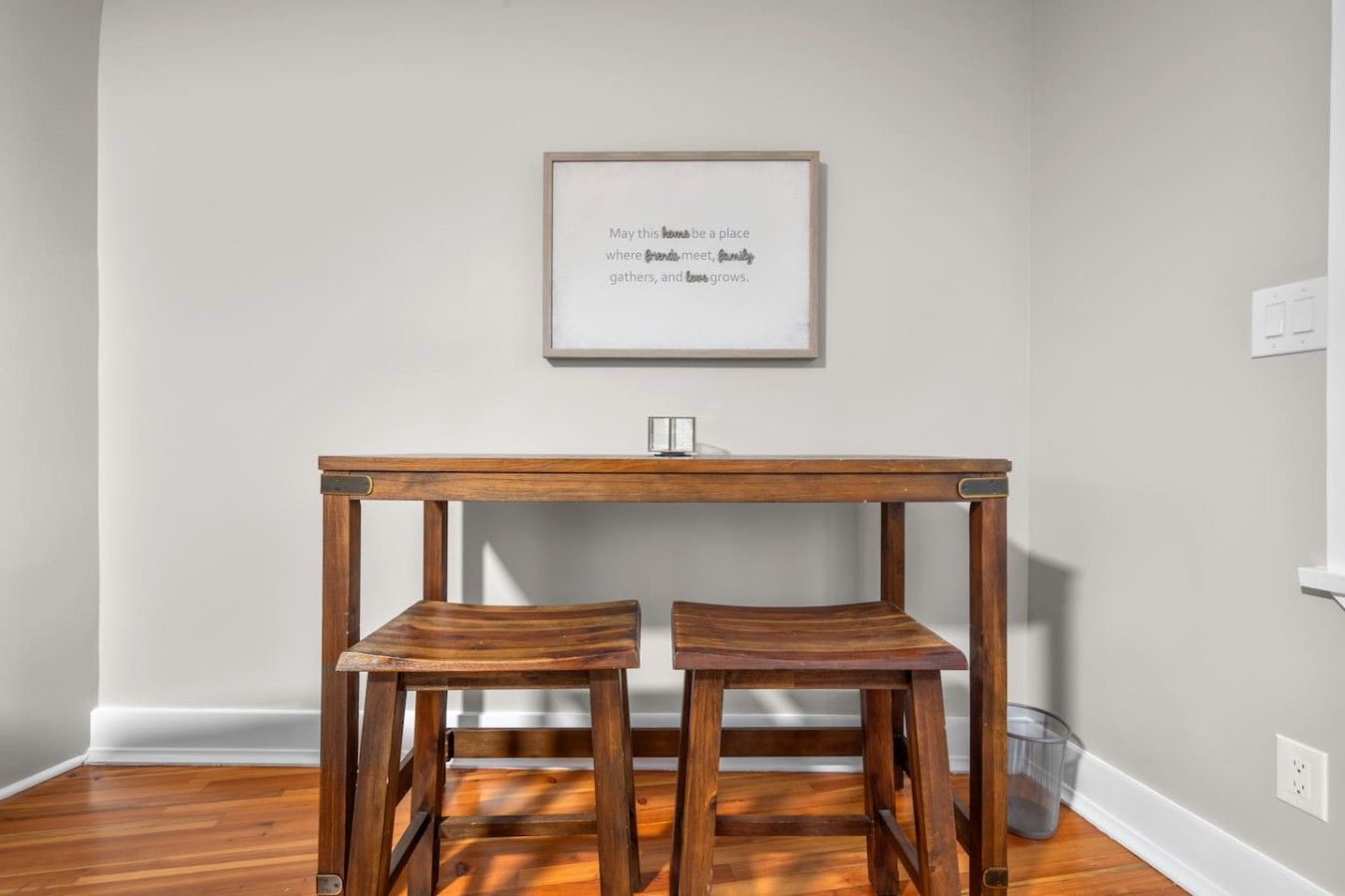 A room with a wooden table and two matching chairs, a framed quote on the wall, and a light switch on the wall.