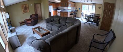 A living room with a large sectional sofa, a coffee table, and a TV stand. There is a dining area with a table and chairs, and a kitchen in the background.