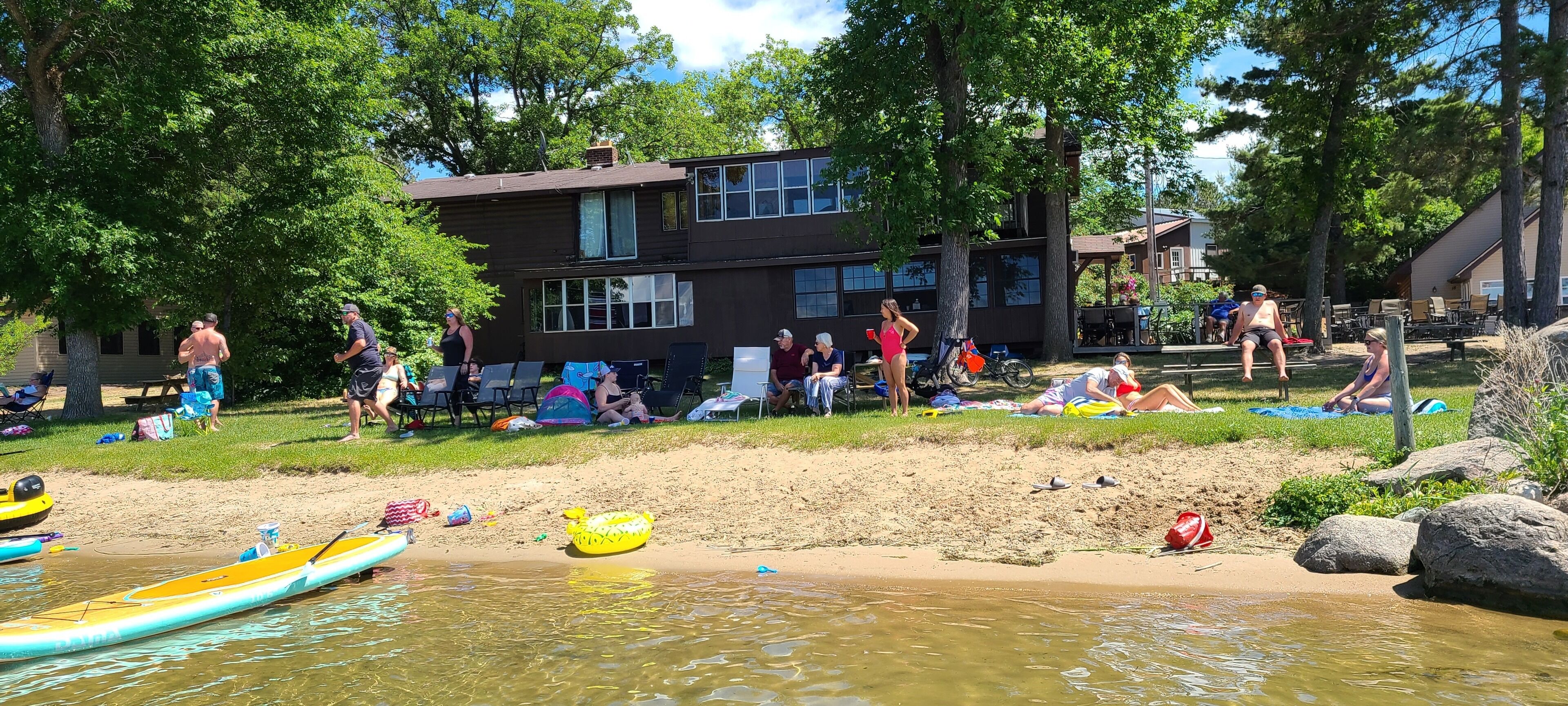 A beach area with people relaxing, a building in the background, and various beach equipment like kayaks and a raft.