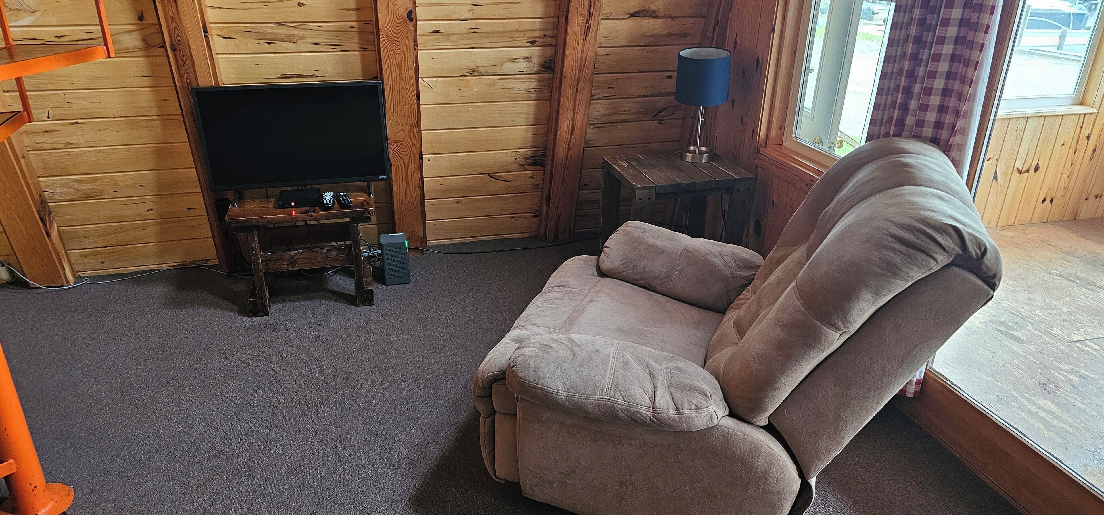 A cozy room with a wooden interior, a brown sofa, a television on a wooden stand, and a lamp on a small table.