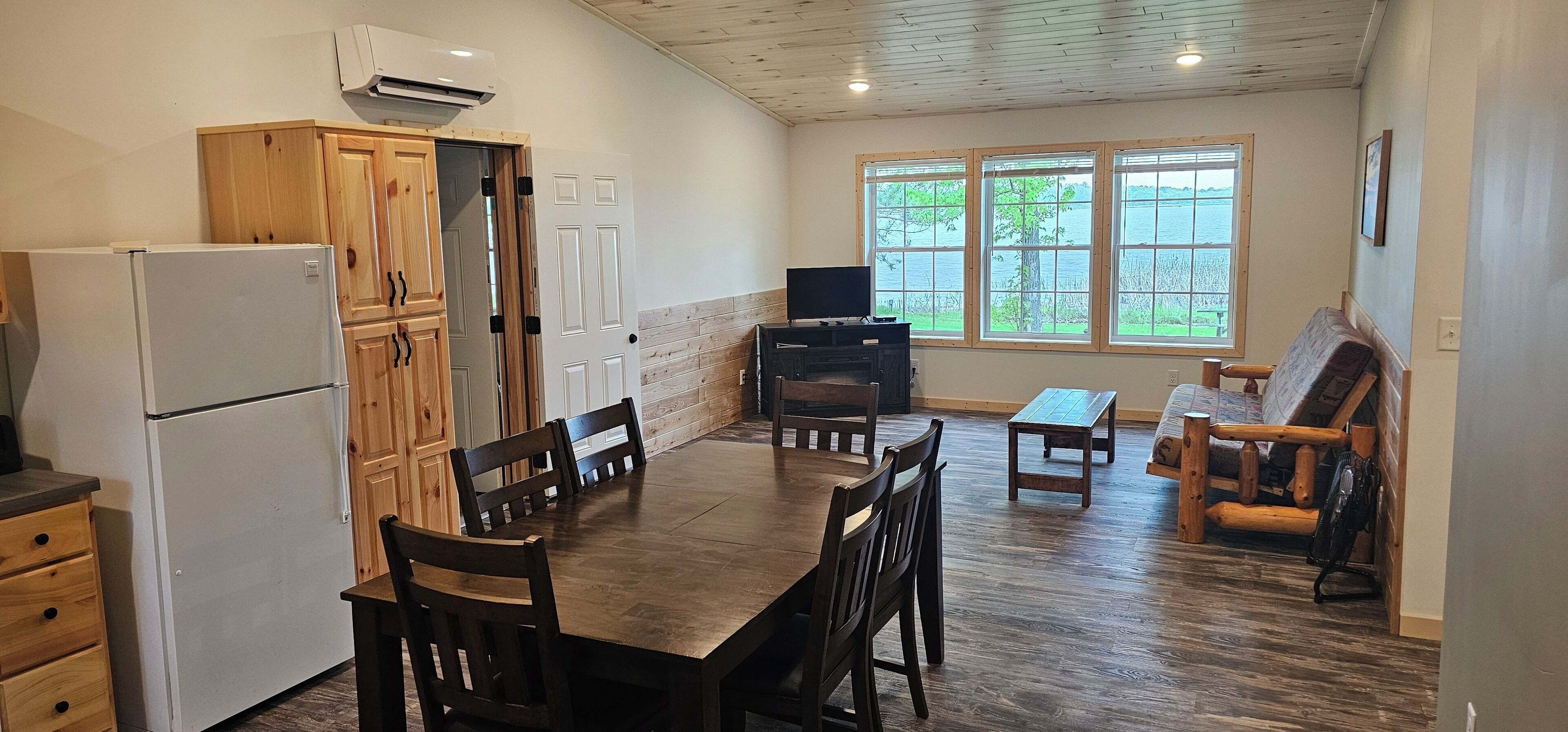 A kitchen with wooden cabinets, a white refrigerator, a dining table with chairs, a television, and a large window with a view of trees and water.