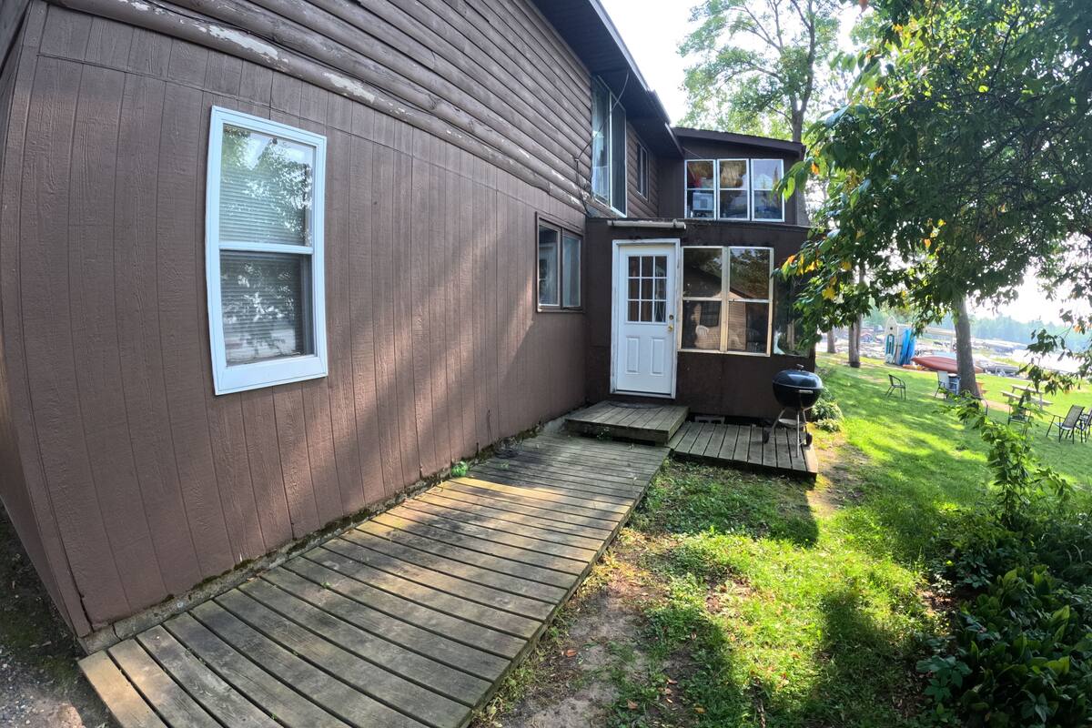 Une maison brune avec une terrasse en bois menant à la porte d’entrée, un barbecue et un petit jardin avec un arbre et de la pelouse.