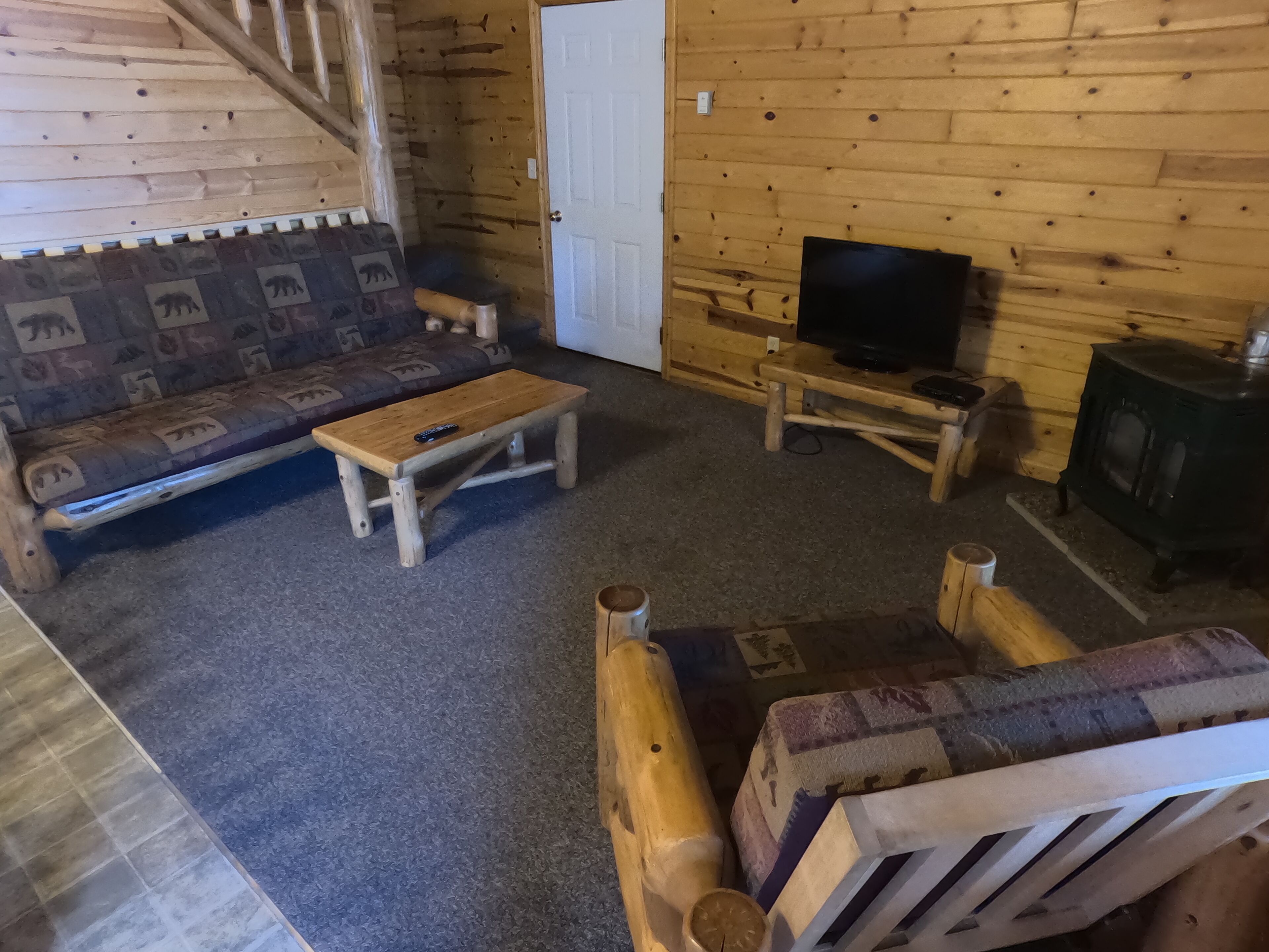 Un acogedor interior de cabaña con una escalera de madera, un sofá, una mesa de centro, un televisor y un estufa de leña.
